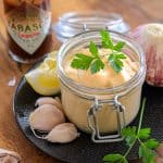A glass jar of chipotle aioli garnished with parsley, set on a black plate with garlic cloves and a lemon wedge.