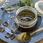 A jar of truffle oil with black truffle slices floating in it, placed on a blue plate with a sprig of fresh green herbs and coarse sea salt sprinkled around.