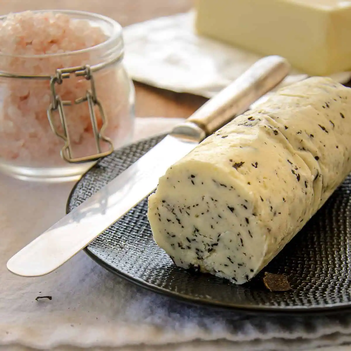 A roll of truffle butter on a plate with a knife and a jar of pink salt beside it.