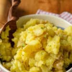 A bowl of roasted garlic mashed potatoes placed on a wooden surface, with a wooden spoon ready for serving, giving a homely and inviting look.