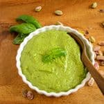 A bowl of vibrant green pistachio pesto garnished with fresh basil leaves, surrounded by scattered pistachios on a wooden surface.
