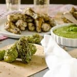 Grilled chicken wings coated in green pesto sauce placed on a wooden board, with a bowl of green pesto dip and a plate of additional chicken wings in the background. A white napkin is laid next to the board.