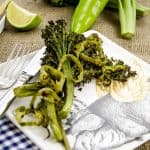 A serving of roasted baby broccolini on a decorative plate, with a fork and knife placed beside it, surrounded by fresh lime and green peppers.
