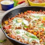 A cast iron skillet filled with a colorful chorizo and eggs dish, garnished with chopped cilantro and surrounded by fresh ingredients in the background.