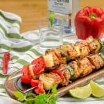 Grilled Southwestern-style pork kabobs with red bell pepper, onion, and zucchini, served on a wooden platter with lime wedges and a bottle of tequila in the background.