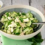 A bowl of cucumber feta salad, garnished with fresh mint leaves and finely sliced shallots. The salad is set on a green napkin, with a rustic wooden background.
