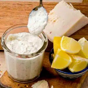 A close-up of a jar of garlic Parmesan wing sauce with a spoonful of sauce above it, lemon wedges, and a block of Parmesan cheese on a wooden board.