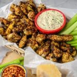 A tray of Italian-seasoned chicken wings served with celery sticks and a bowl of creamy dip, garnished with parsley, and placed on parchment paper.