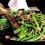 A skillet filled with vibrant green beans, mushrooms, and bacon bits, cooked to perfection. A wooden spoon is placed in the skillet, and a white towel with a red checkered pattern is draped over the handle.