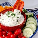 Keto cream cheese dip in a red bowl, garnished with parsley, with a vibrant assortment of fresh vegetables like cherry tomatoes, cucumber slices, and bell pepper slices around it on a blue and white checkered cloth.