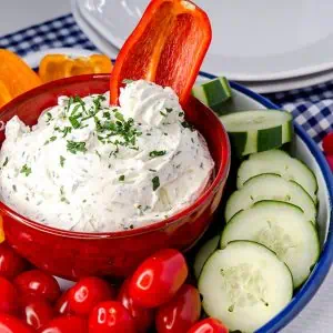 Keto cream cheese dip in a red bowl, garnished with parsley, with a vibrant assortment of fresh vegetables like cherry tomatoes, cucumber slices, and bell pepper slices around it on a blue and white checkered cloth.