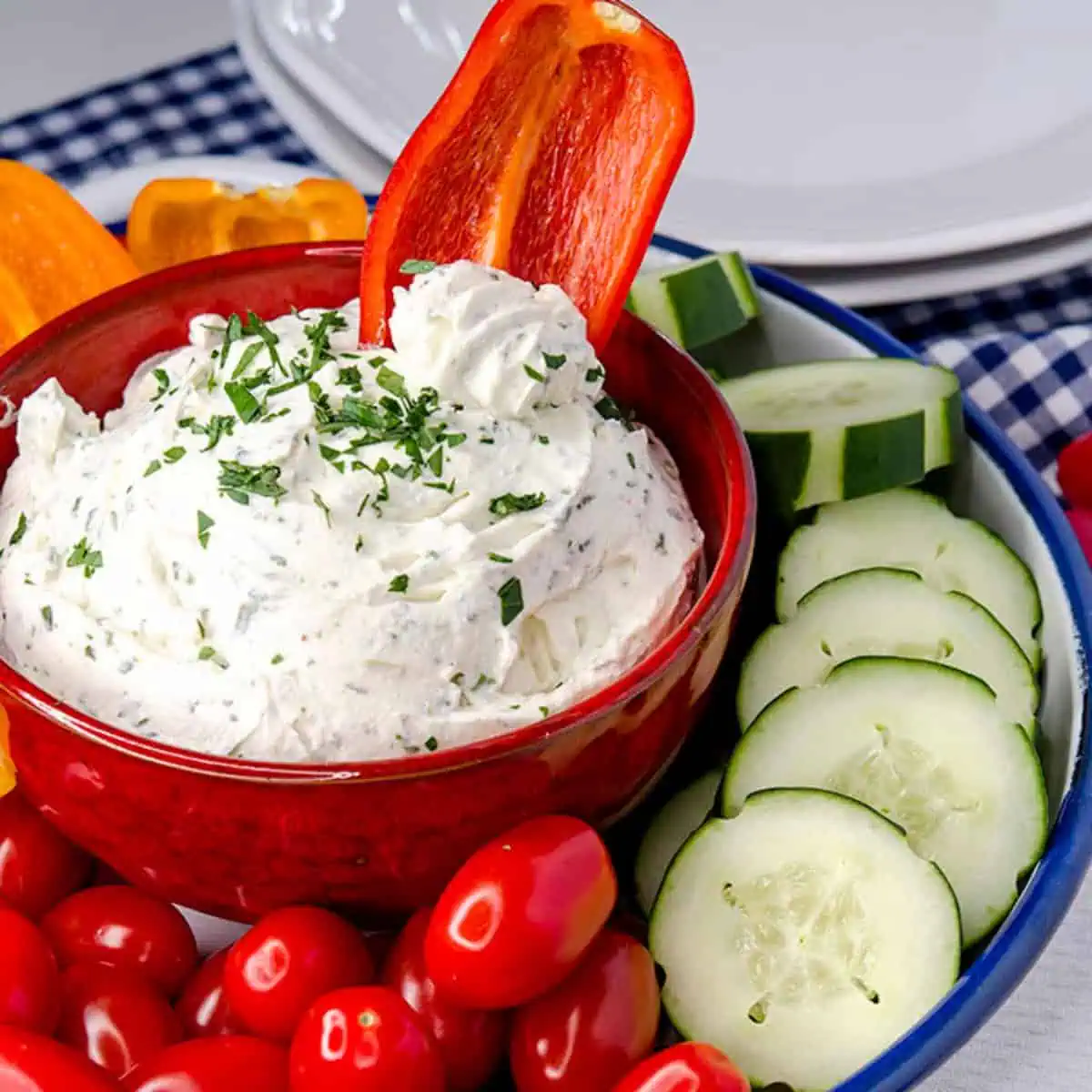 Keto cream cheese dip in a red bowl, garnished with parsley, with a vibrant assortment of fresh vegetables like cherry tomatoes, cucumber slices, and bell pepper slices around it on a blue and white checkered cloth.