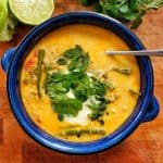 An overhead view of a bowl of Thai red curry soup garnished with fresh cilantro, with lime wedges on the side, set on a wooden cutting board.