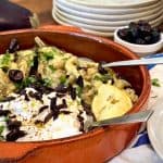 A close-up of a hand holding a cucumber slice topped with a roasted eggplant mixture, Greek yogurt, and an olive, with a ceramic dish of the appetizer in the background.