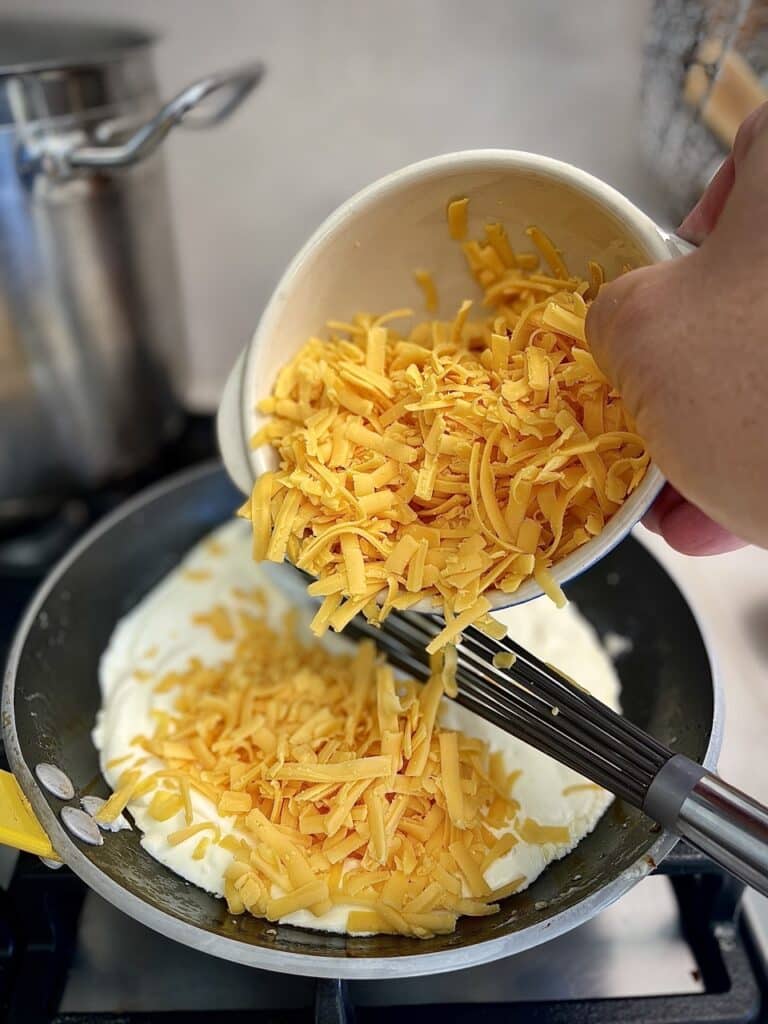 Adding shredded cheddar cheese to the melted cream cheese in a pan.