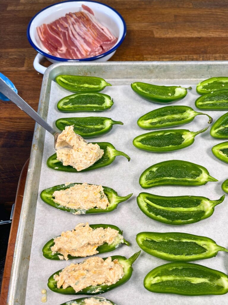 Mixture of ingredients being added into halved seedless jalapeños on a baking tray