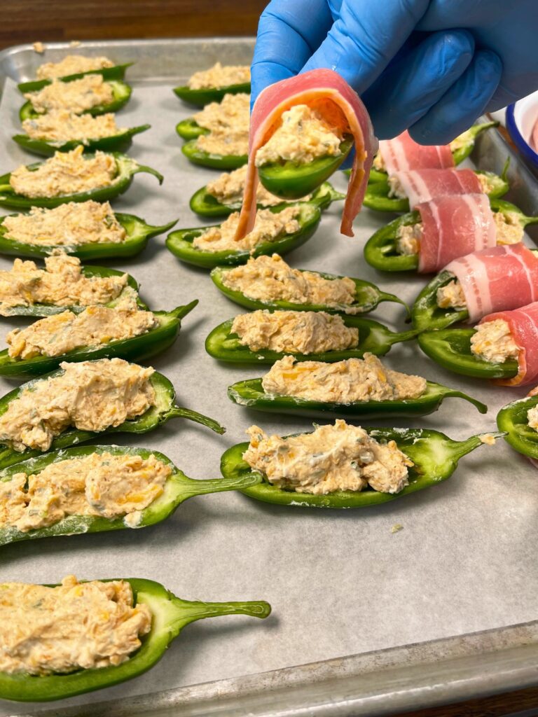 Bacon being wrapped around filled halved jalapeños