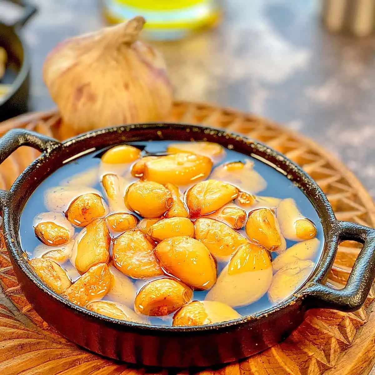 A small cast iron pan filled with golden, slow-roasted garlic cloves, glistening in oil, set on a wooden serving board.