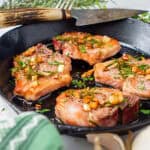 Lamb loin chops cooked to perfection in a cast iron skillet, garnished with garlic and rosemary, with a green towel and garlic bulb nearby.