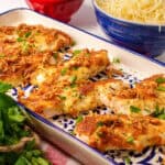 A tray of golden, cheese-crusted Keto Chicken Tenders garnished with fresh parsley. Bowls of shredded cheese and fresh parsley are visible in the background, enhancing the presentation.