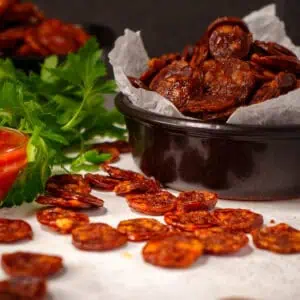 A black ceramic bowl filled with crispy, golden-brown pepperoni chips, with a few scattered around the bowl on a white parchment paper. Fresh parsley and a small bowl of marinara sauce are in the background, adding a vibrant green and red contrast.