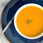 A bowl of vibrant Butternut Squash soup garnished with a small sprig of fresh thyme. The bowl is set on a dark plate with a silver spoon beside it, and the background includes a soft white cloth, creating a warm and inviting presentation.