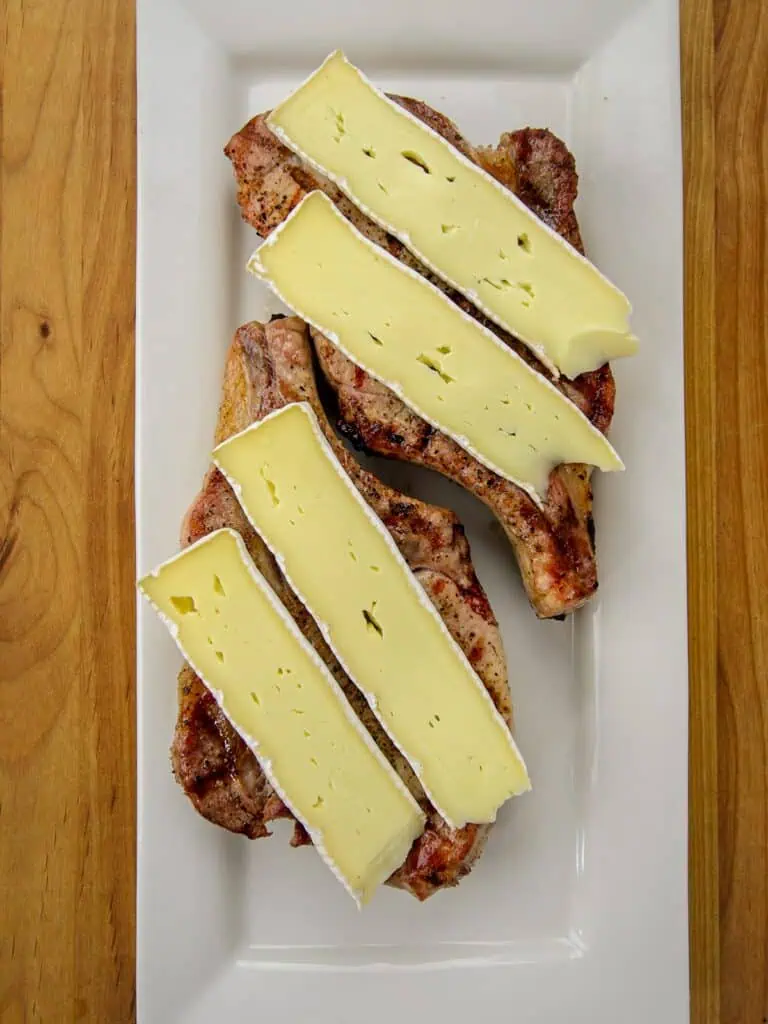 Grilled pork chops topped with slices of brie cheese on a white rectangular plate.