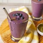 A close-up view of a purple protein shake in a glass with a metal straw, topped with blueberries and a sprinkle of cinnamon, placed on a wooden board with a yellow and white checkered napkin.