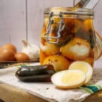 A glass jar filled with spicy pickled eggs, surrounded by fresh ingredients like jalapeno peppers, garlic, and eggs on a wooden cutting board.