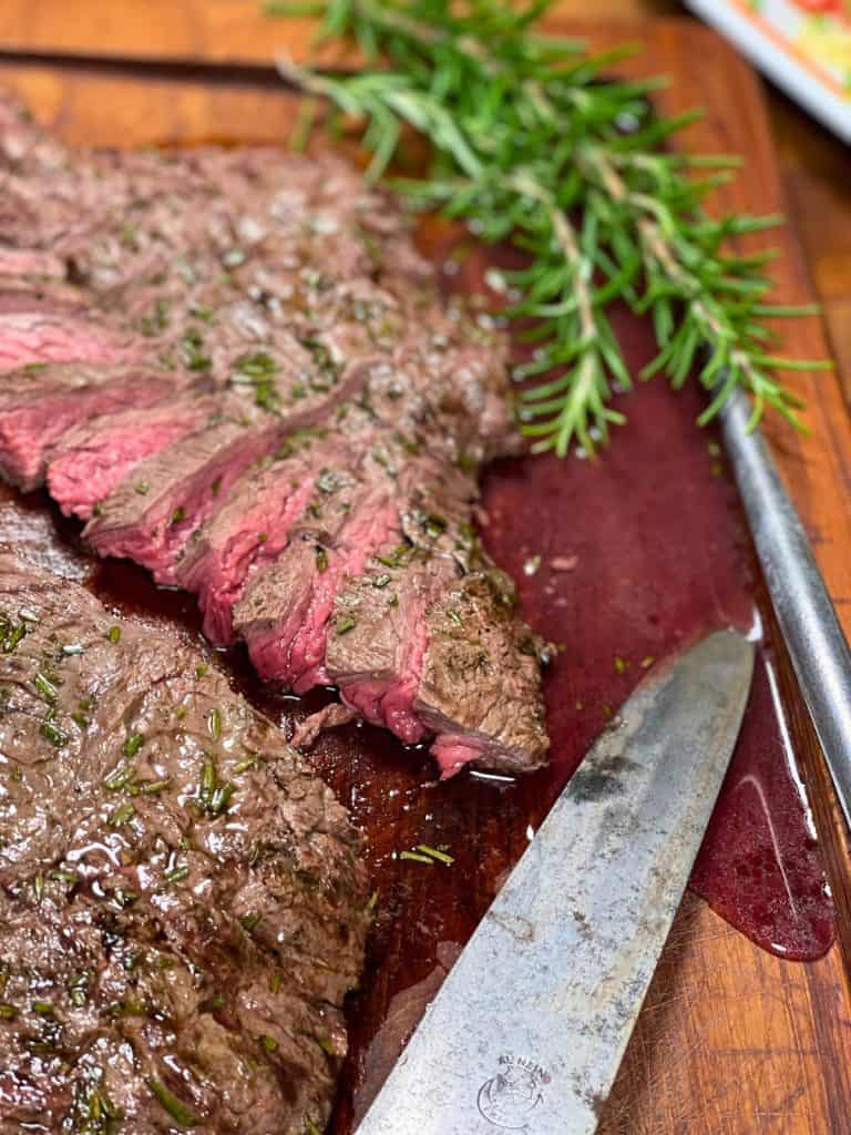 Sliced steak on a wooden cutting board, garnished with rosemary and garlic, with a vintage knife and a meat fork nearby.