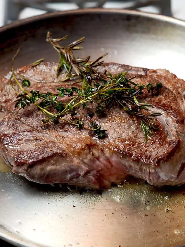A close-up of a cooked beef chuck roast garnished with fresh rosemary and thyme, with red onions and garlic cloves in the background.