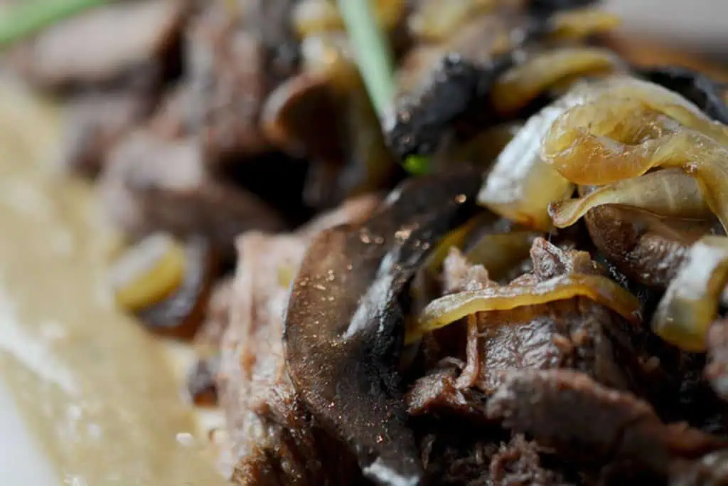 A close-up of a beef chuck roast with caramelized onions and mushrooms, garnished with fresh herbs, served on a white plate.