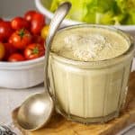 A close-up of a glass jar filled with Caesar salad dressing, garnished with grated Parmesan cheese, with a spoon leaning against the jar. In the background, there are bowls of cherry tomatoes and lettuce, creating a fresh and vibrant setting.