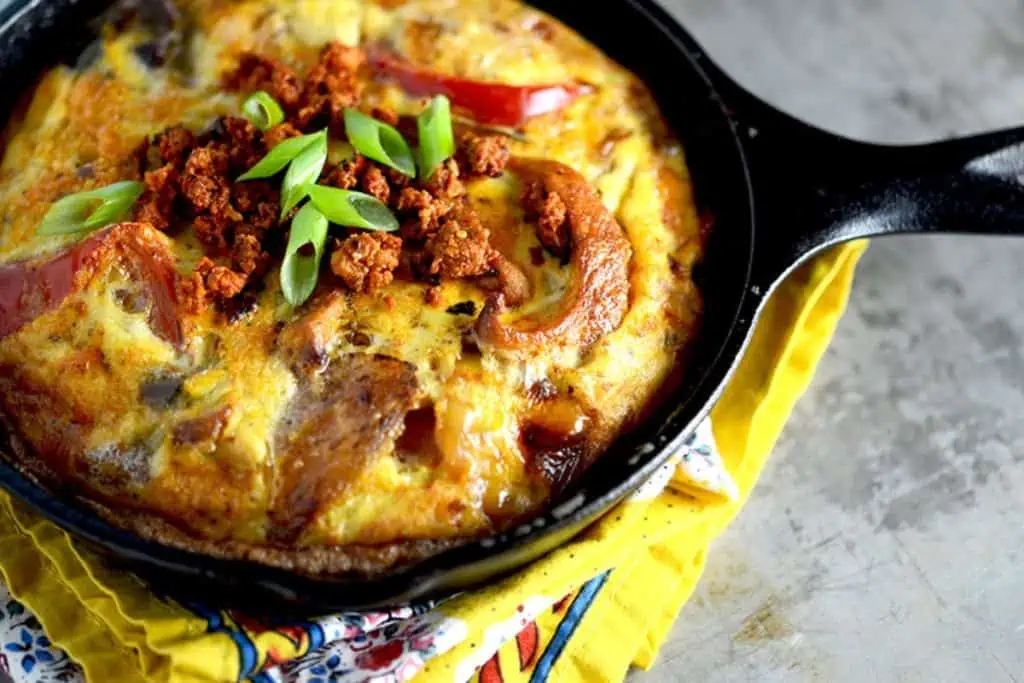 A top view of an Easy Fajita Frittata cooked in a cast iron skillet, featuring visible chunks of chorizo, red bell peppers, and green onions on top, with a yellow napkin underneath the skillet.