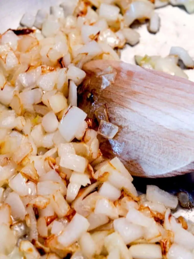 A close-up view of diced onions sautéing in a pan, turning golden brown, with a wooden spoon stirring them.