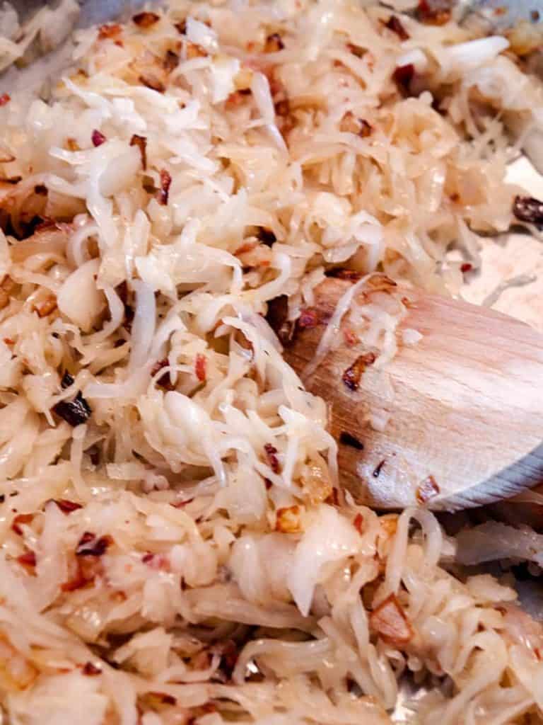 Raw sauerkraut being mixed with sautéed onions and bacon in a pan, stirred by a wooden spoon.