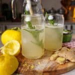 A close-up of two glasses of ginger lemonade with rosemary garnishes, set on a wooden board with fresh lemons, ginger, and lemon zest scattered around. A glass pitcher of lemonade is visible in the background.