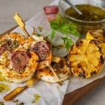 Grilled shrimp skewers with slices of Italian sausage, garnished with herbs and oil, accompanied by grilled lemon halves and a bowl of dipping sauce.