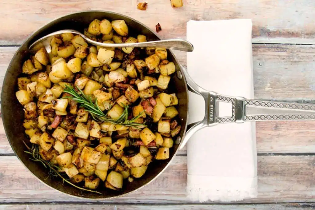 Overhead view of a skillet filled with homemade hash browns and bacon, garnished with rosemary, on a wooden table with a white napkin.