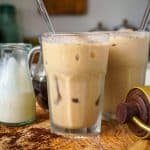 A glass of iced coffee latte with a clear straw, placed on a wooden board. The latte is a light brown color, and the glass is surrounded by pitchers of cream and coffee concentrate, with coffee grounds scattered on the board.