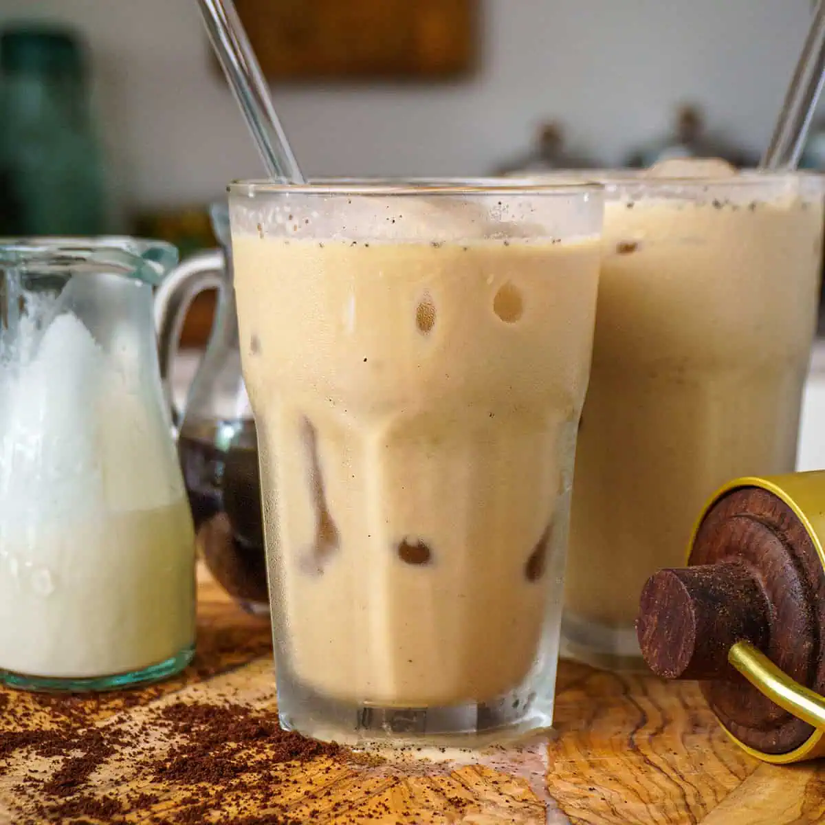 A glass of iced coffee latte with a clear straw, placed on a wooden board. The latte is a light brown color, and the glass is surrounded by pitchers of cream and coffee concentrate, with coffee grounds scattered on the board.
