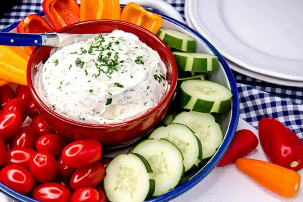 A bowl of creamy keto cream cheese dip garnished with chopped herbs, surrounded by fresh vegetables like sliced cucumbers, cherry tomatoes, and bell pepper strips, displayed on a blue and white checkered tablecloth.