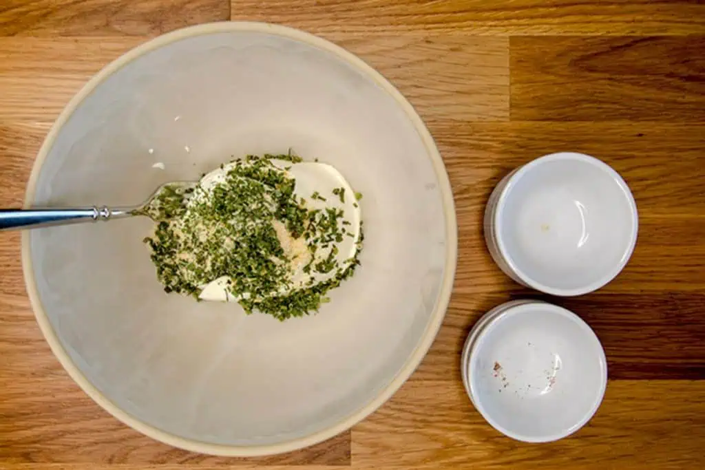 A bowl with the ingredients for keto cream cheese dip, including cream cheese and dried herbs, being mixed together with a fork.