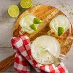 Top view of three glass mugs filled with ice and limeade, garnished with lime wedges, placed on a wooden board with a red and white checkered cloth.