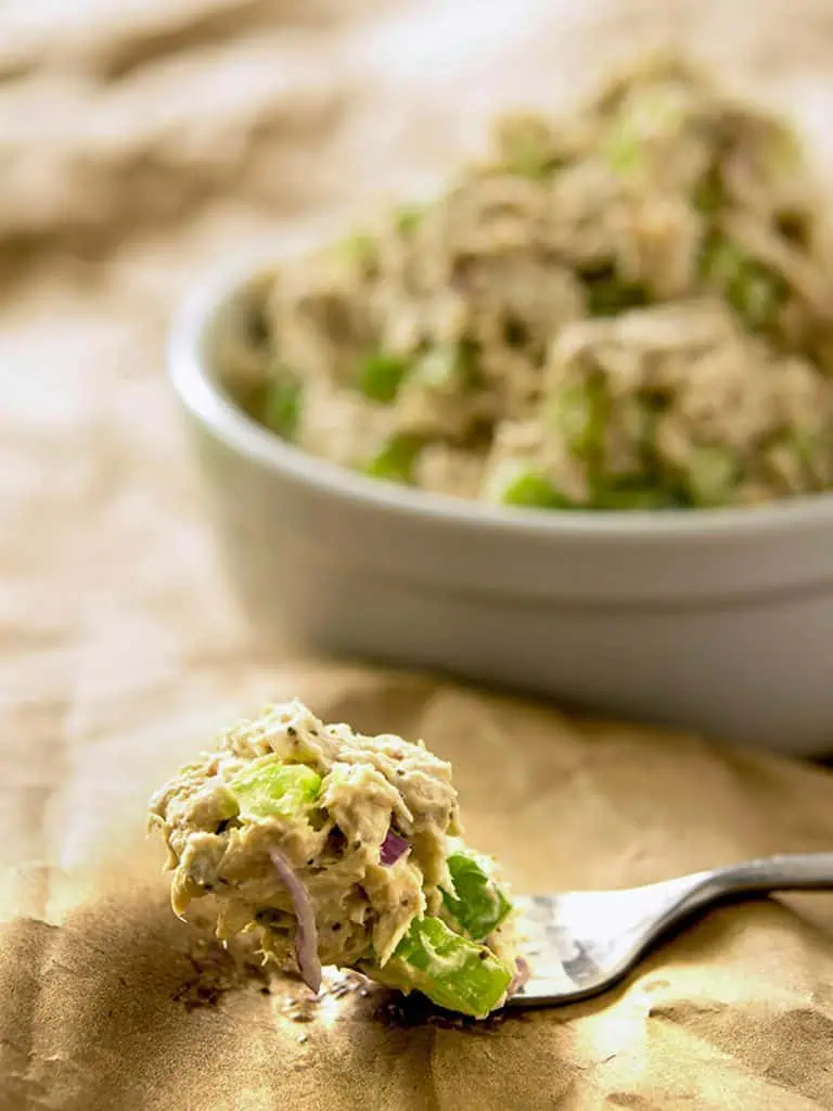 Close-up of a forkful of low-carb tuna salad with visible chunks of tuna, diced celery, and red onion. The fork is on brown parchment paper, with a bowl of the same salad blurred in the background.