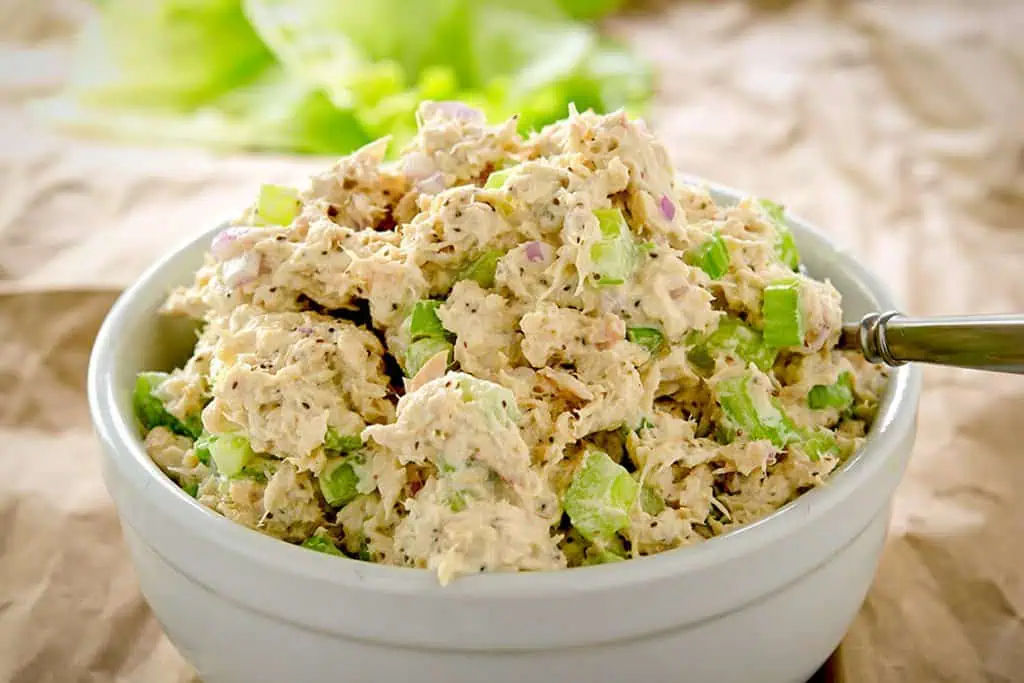 A white bowl filled with low-carb tuna salad, featuring chunks of tuna, diced celery, and red onion. The bowl is placed on a crumpled brown parchment paper.