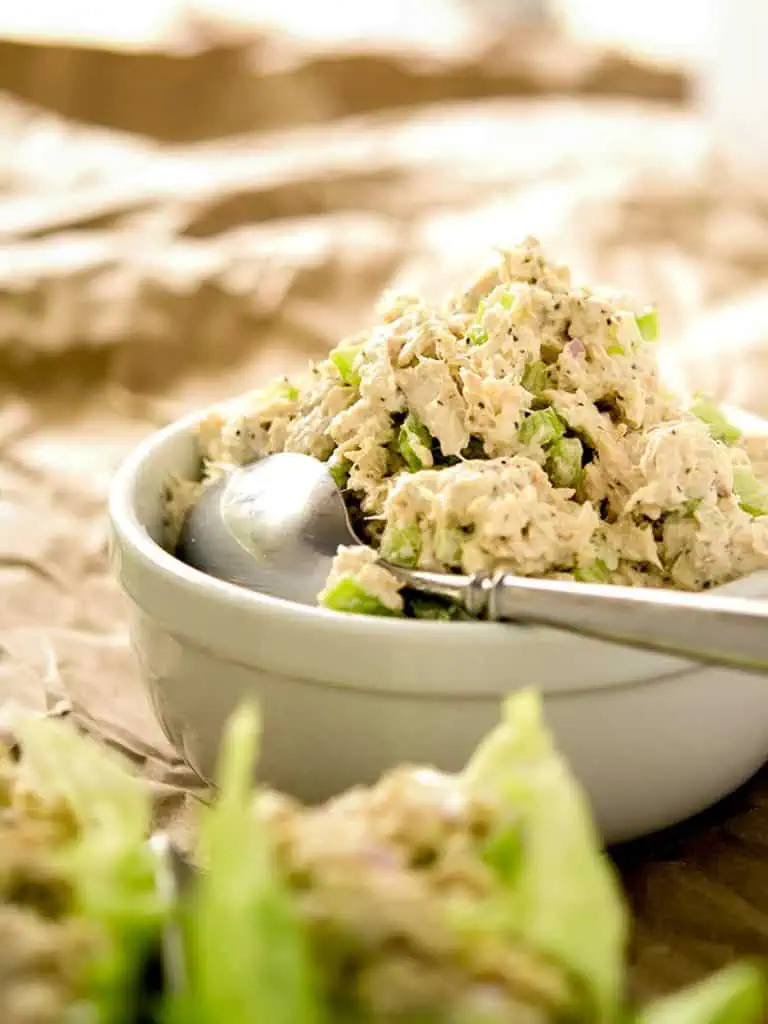 Overhead view of a white bowl filled with low-carb tuna salad, showcasing chunks of tuna, diced celery, and red onion. The bowl is placed on crumpled brown parchment paper with green lettuce in the background.