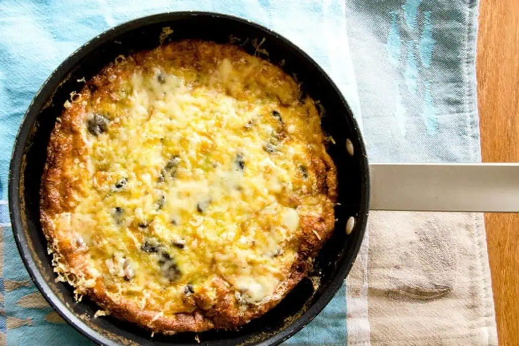 An overhead view of a golden-brown mushroom and leek frittata in a black pan.