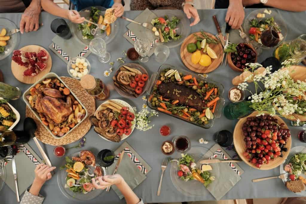 Overhead view of a festive table spread with various dishes, including a whole roasted chicken, roasted vegetables, fresh fruits, and garnished with flowers. People are seated around the table, eating and enjoying the meal.