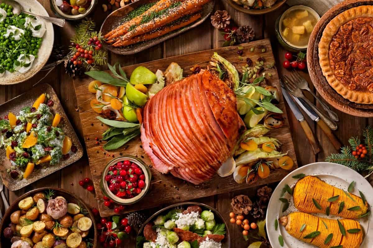 A beautifully arranged holiday feast featuring a large honey-glazed ham as the centerpiece. Surrounding the ham are various side dishes like roasted vegetables, salads, and a pie, all set on a wooden table decorated with holiday-themed garnishes.